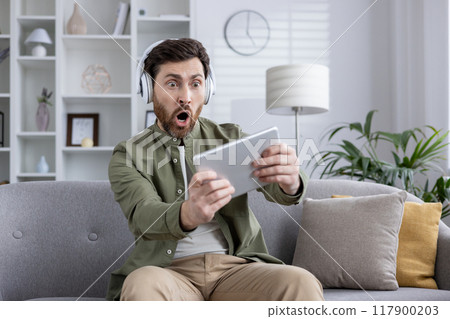 Man sitting on couch wearing headphones, using tablet for video game. Expresses excitement and surprise, likely from gaming experience. Reflects themes of technology, entertainment, modern lifestyle. 117900203