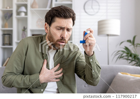 Man holding asthma inhaler, appearing worried and placing hand on chest. Seated on sofa, managing respiratory issues. Indoor setting suggests home environment, highlighting personal health care 117900217