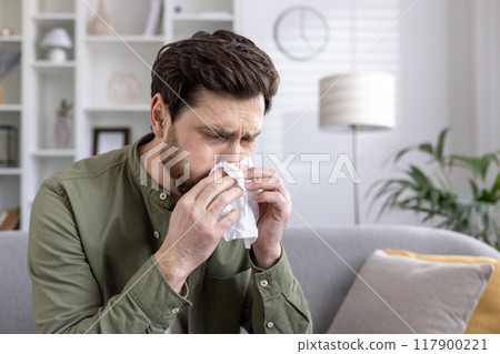 Man sneezing into tissue on couch shows discomfort from allergies or cold. Indoor setting with cozy atmosphere. Scene depicts health, illness, home remedies, and seasonal issues. 117900221