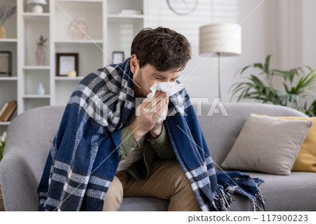 Man sitting on couch wrapped in a blue blanket, sneezing into tissue. Scene depicts illness, cold, flu symptoms, and seeking comfort at home. Expression of discomfort and need for care. 117900223