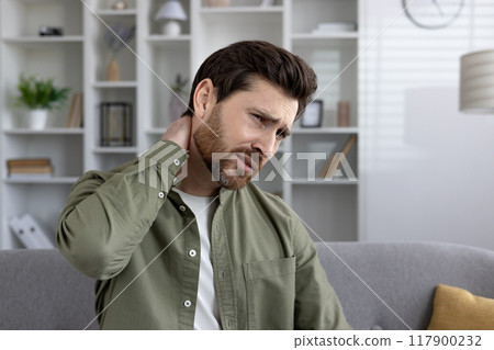 Man wearing green shirt holds neck with expression of pain, sitting on sofa. Experiencing discomfort in home setting, showcasing feelings of stress and tension. Man wearing green shirt holds neck with expression of pain, sitting on sofa. Experiencing discomfort in home setting, showcasing feelings of stress and tension. 117900232