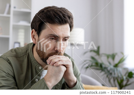Pensive man in casual clothing sits thoughtfully at home, hands clasped near face. Capturing mood of contemplation and introspection, perfect for themes of personal reflection, decisions, thinking. 117900241