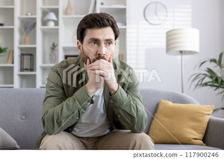 Thoughtful man in casual clothes sits on couch in modern living room, lost in contemplation. Calm atmosphere signifies introspection, choice-making. Emotion conveyed is pensive, thoughtful reflection Thoughtful man in casual clothes sits on couch in modern living room, lost in contemplation. Calm atmosphere signifies introspection, choice-making. Emotion conveyed is pensive, thoughtful reflection 117900246