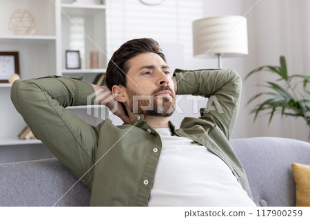 Relaxed man sits on couch with hands behind head, reflecting in modern living room. Casual attire complements calm demeanor, conveying tranquility and peacefulness. Relaxation and contemplation 117900259