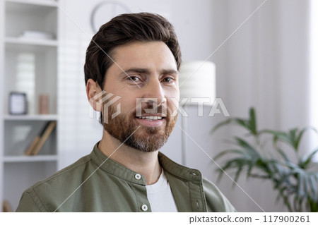 Smiling man with beard wearing casual shirt in modern office environment. Confident expression and casual attire suggest professional or creative workplace. 117900261