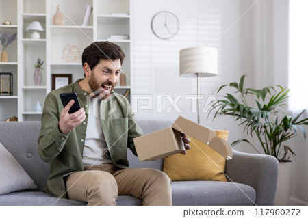 Angry male buyer received damaged parcel, man sitting on sofa in living room with phone in hands and open cardboard box. 117900272