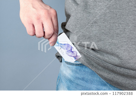 A young man takes out a credit card from his blue jeans pocket. Close-up on a hand taking out a payment card from trousers on a light grey studio background. Business and payment concept A young man takes out a credit card from his blue jeans pocket. Close-up on a hand taking out a payment card from trousers on a light grey studio background. Business and payment concept 117902388
