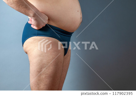 Side view of a fat man with a protruding belly wearing blue underwear on a grey background. Studio portrait for health and fitness concepts Side view of a fat man with a protruding belly wearing blue underwear on a grey background. Studio portrait for health and fitness concepts 117902395