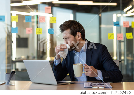 Businessman in suit sneezing at desk while using laptop, holding coffee mug. Represents health issues, cold, flu, and work challenges. Highlights professional setting, stress, and well-being office. 117902490