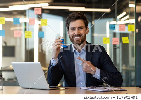 Smiling businessman using inhaler for asthma while working in modern office. Man in suit pointing at inhaler next to laptop and notebook. Positive expression, health concept, work-life balance. 117902491