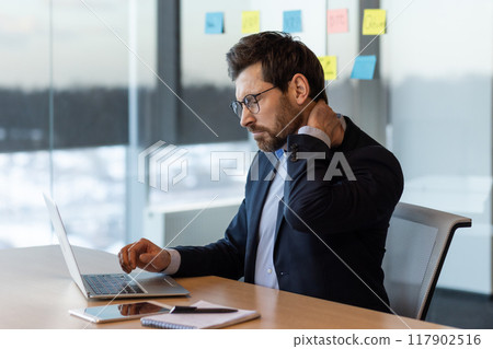 Businessman in suit sitting at desk using laptop, appearing uncomfortable due to neck pain. Office with focused work atmosphere illustrates concept of work-related stress and physical discomfort Businessman in suit sitting at desk using laptop, appearing uncomfortable due to neck pain. Office with focused work atmosphere illustrates concept of work-related stress and physical discomfort 117902516