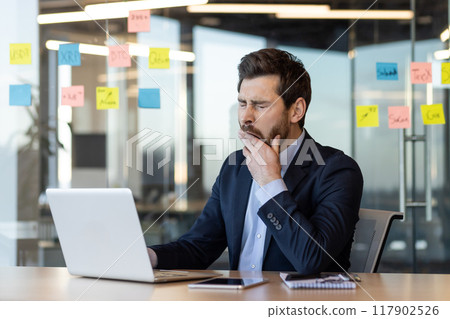 Exhausted businessman wearing suit working on laptop in office yawning with hand covering mouth. Modern work environment with sticky notes. Concept of fatigue, tiredness, and office stress. 117902526