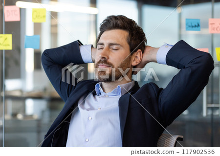 Businessman relaxing at office desk with hands behind head, eyes closed. Enjoying moment of peace and calm, taking break from work, wearing a formal suit. 117902536