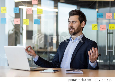 Businessman practicing meditation in office for relaxation and stress relief. Man sitting at desk with laptop , maintaining work-life balance and mindfulness 117902540