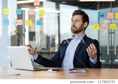 Businessman practicing meditation at desk surrounded by sticky notes and laptop. Focus on stress relief, mindfulness, and mental clarity in busy office environment. Relaxation techniques in workplace 117902541