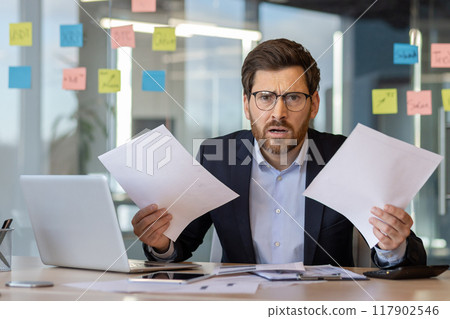 Businessman at office desk overwhelmed by work holding papers. Frustrated expression suggests stress and urgency. Laptop and phone on desk indicate multitasking in modern workplace setting. 117902546