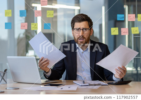 Businessman in suit sitting at desk with documents, expressing confusion. Laptop and phone on table suggest analysis of data. Office setting with notes on glass wall, indicating problem-solving. 117902557