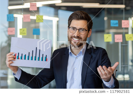 Professional businessman confidently presenting data analysis using colorful bar graph. Engaging smile and welcoming gesture create positive business environment. 117902581