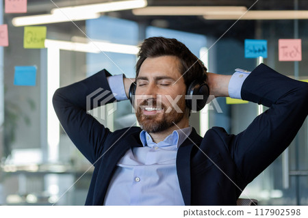 Businessman in office enjoying relaxation with headphones, smiling while listening to music. Professional attire and positive emotions suggest stress-free work environment. Businessman in office enjoying relaxation with headphones, smiling while listening to music. Professional attire and positive emotions suggest stress-free work environment. 117902598