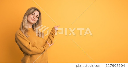 Cheerful young woman showing side space with fingers on yellow background Cheerful young woman showing side space with fingers on yellow background 117902604