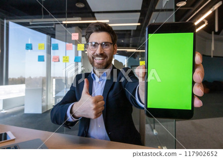 Businessman in modern office shows smartphone with green screen, expressing positivity with thumbs up gesture. Ideal for technology, success, and professional concepts in corporate environment. Businessman in modern office shows smartphone with green screen, expressing positivity with thumbs up gesture. Ideal for technology, success, and professional concepts in corporate environment. 117902652