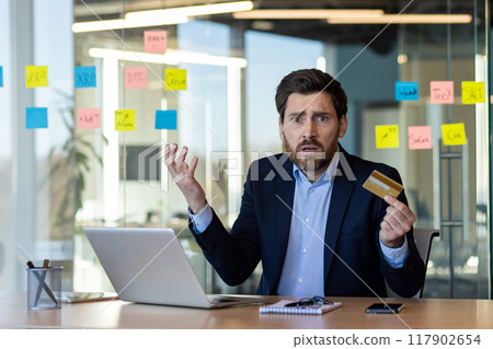 Confused businessman sitting at desk holding credit card showing frustration at laptop. Professional in suit working in modern office surrounded by notes. Concept of stress, business problem, online Confused businessman sitting at desk holding credit card showing frustration at laptop. Professional in suit working in modern office surrounded by notes. Concept of stress, business problem, online 117902654
