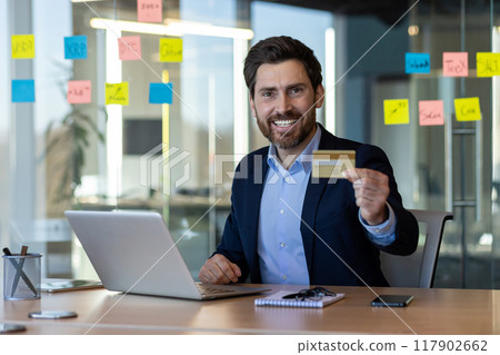 Confident businessman in formal attire holding credit card at office desk, working on laptop, showing happiness and satisfaction. Colorful sticky notes on glass wall suggesting planning or Confident businessman in formal attire holding credit card at office desk, working on laptop, showing happiness and satisfaction. Colorful sticky notes on glass wall suggesting planning or 117902662