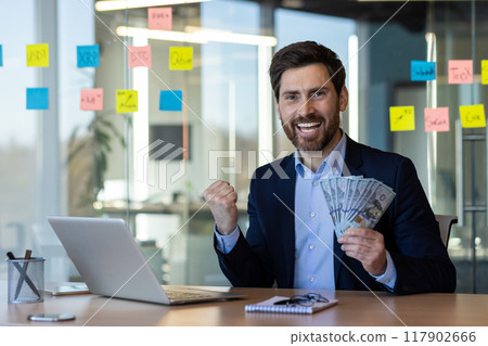 Businessman celebrating financial success holding cash at desk with laptop and office supplies. 117902666