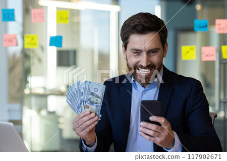Businessman expresses happiness holding cash and checking smartphone. Man dressed in formal suit shows achievement and financial success, depicting concepts of wealth, communication, and business. Businessman expresses happiness holding cash and checking smartphone. Man dressed in formal suit shows achievement and financial success, depicting concepts of wealth, communication, and business. 117902671