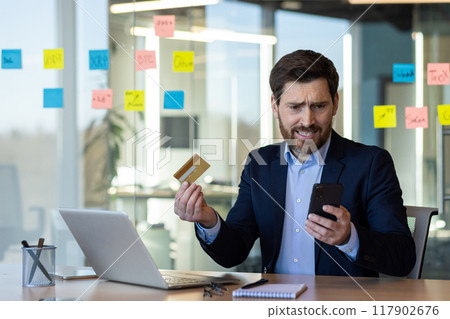 Businessman holds credit card with worried expression while checking smartphone in modern office. Indicates concern about online banking, technology issues, or payment problems. Businessman holds credit card with worried expression while checking smartphone in modern office. Indicates concern about online banking, technology issues, or payment problems. 117902676