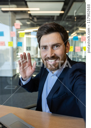 Smiling businessman waving hand during video call in office. Professional wearing suit communicating through phone. Modern workplace with sticky notes on glass walls Smiling businessman waving hand during video call in office. Professional wearing suit communicating through phone. Modern workplace with sticky notes on glass walls 117902688