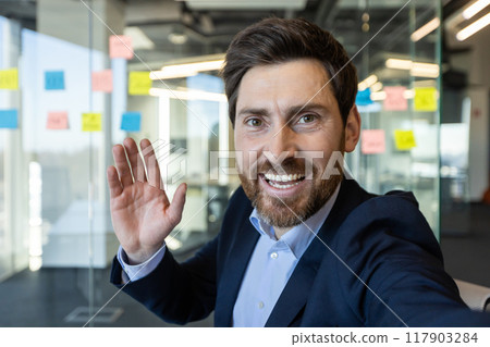 Smiling businessman waving during video call using phone in modern office. Professional appearance, friendly gesture, collaborative ambiance. 117903284