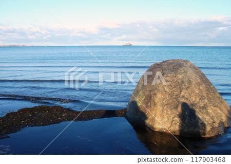 Solitary rock stands in shallows of calm sea, with distant ship on horizon. Seascape with large boulder in the foreground and a vessel in distance. Tranquil beach scene featuring rock and ocean Solitary rock stands in shallows of calm sea, with distant ship on horizon. Seascape with large boulder in the foreground and a vessel in distance. Tranquil beach scene featuring rock and ocean 117903468