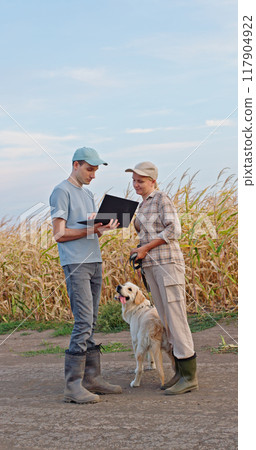 Two farmers using a laptop in a cornfield, with a Golden Retriever standing beside them. Two farmers using a laptop in a cornfield, with a Golden Retriever standing beside them. 117904922