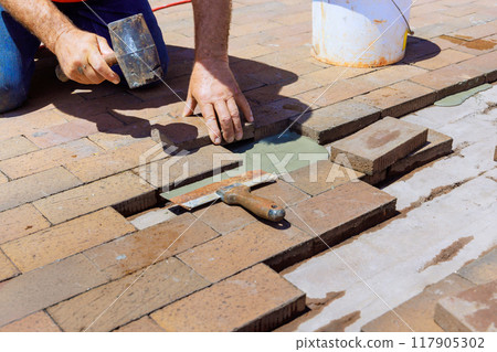 An employee is engaged in repairing footpath by replacing damaged bricks during reconstruction 117905302