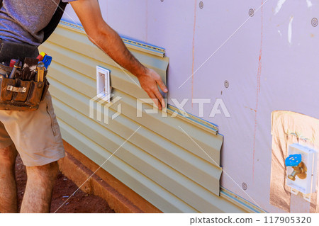 An employee installs vinyl plastic siding panels on wall facade of new residence 117905320