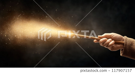 A hand holding a wooden wand, emitting glowing golden magic against a dark background 117907157