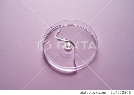 Serum or cosmetic oil flows into a transparent bowl on a purple background. 117910868