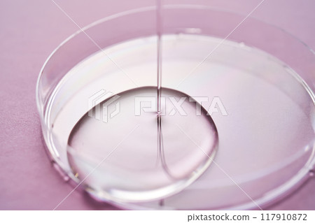 Serum or cosmetic oil flows into a transparent bowl on a purple background. Serum or cosmetic oil flows into a transparent bowl on a purple background. 117910872