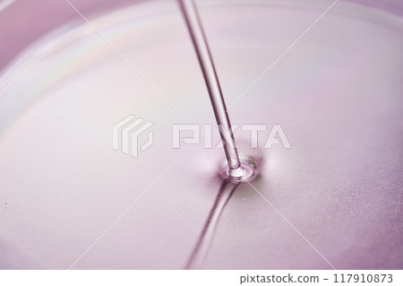 Serum or cosmetic oil flows into a transparent bowl on a purple background. Serum or cosmetic oil flows into a transparent bowl on a purple background. 117910873