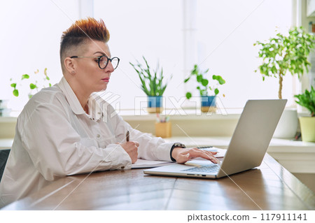 Middle-aged serious woman working at computer laptop in home office Middle-aged serious woman working at computer laptop in home office 117911141