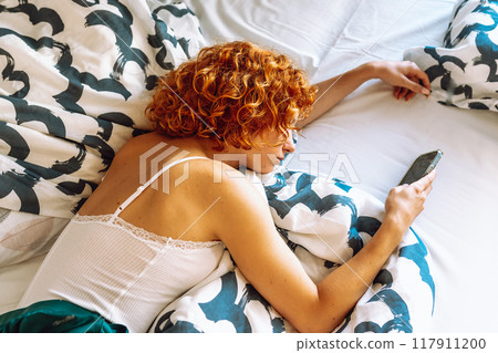 portrait of red-haired young woman lying on bed using mobile phone 117911200