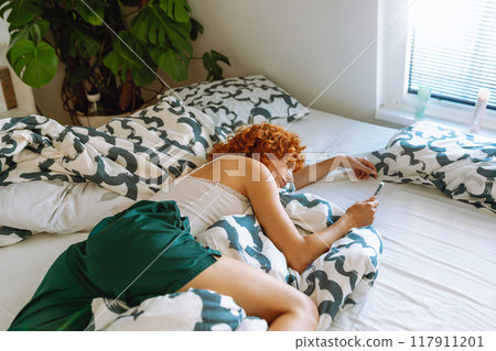 portrait of red-haired young woman lying on bed using mobile phone 117911201