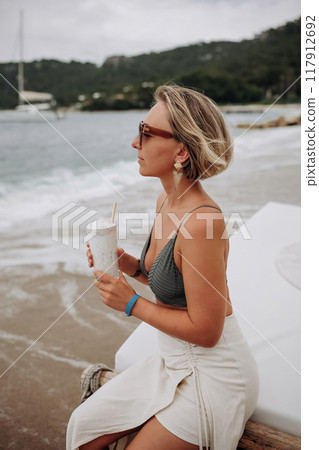 A woman enjoying a refreshing drink on the beach while watching the waves on a cloudy day A woman enjoying a refreshing drink on the beach while watching the waves on a cloudy day 117912692