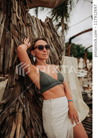 A woman in a stylish bikini and skirt relaxes against a wooden structure at a beach resort  117912697
