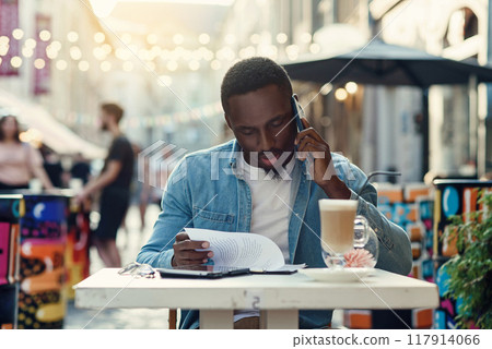 African american business man reads documents and speaking on smartphone sitting on outdoor cafe African american business man reads documents and speaking on smartphone sitting on outdoor cafe 117914066