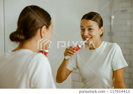 A cheerful and confident woman smiling while brushing her teeth in a contemporary bathroom 117914413