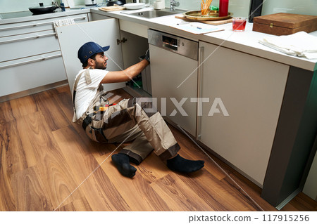 A professional male plumber in uniform is repairing a kitchen sink at home 117915256
