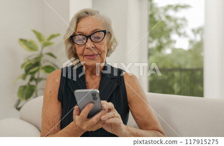 An Elderly Woman Actively Using a Smartphone While Sitting Comfortably in a Modern Living Room 117915275