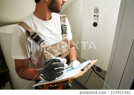 Professional male handyman in uniform is checking gas boiler at home and making notes in tablet Professional male handyman in uniform is checking gas boiler at home and making notes in tablet 117915301